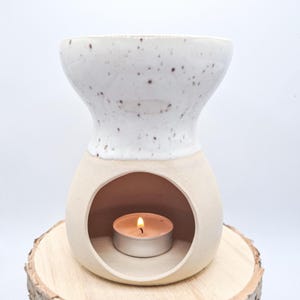 Könnte beinhalten: Ein weißer Keramik-Öldiffusor mit braunen Flecken. Der Diffusor hat eine runde Öffnung am Boden für ein Teelicht. Ein Teelicht befindet sich in der Öffnung und ist angezündet.