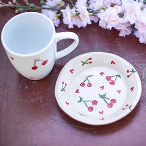 Könnte beinhalten: Weiße Keramiktasse und Untertasse mit Kirschendesign. Die Tasse hat einen Henkel und die Untertasse einen kleinen Rand. Beide Teile sind mit roten Kirschen und grünen Blättern verziert.