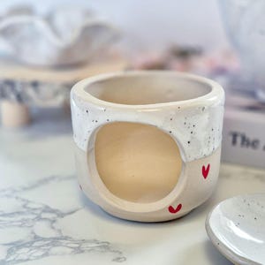 May include: A ceramic mug with a large circular cutout on the side. The upper portion features a white glaze with black speckles, while the lower portion is a smooth beige clay decorated with small, hand-painted red heart shapes.