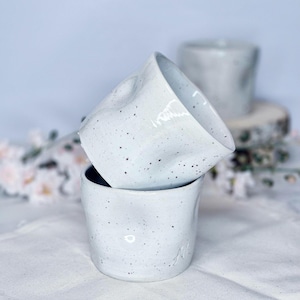 Könnte beinhalten: Zwei weiße Keramikbecher mit gesprenkeltem Design sind übereinander gestapelt. Die Becher haben eine strukturierte, organische Form und ein mattes Finish. Der Hintergrund ist ein weiches, verschwommenes Weiß.