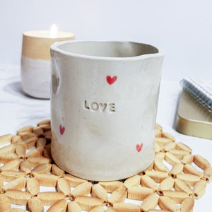 Könnte beinhalten: Eine weiße Keramiktasse mit dem Wort "LOVE" in Relief auf der Vorderseite. Die Tasse ist mit kleinen roten Herzen verziert und steht auf einem geflochtenen Bambusuntersetzer.