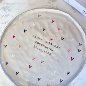 May include: A round, light gray ceramic plate with a wavy, uneven edge. The surface is decorated with small, multi-colored heart icons in red, blue, orange, green, and white. The text "HAPPY BIRTHDAY CONSTANTIN 20.09.2025" is stamped in the center.