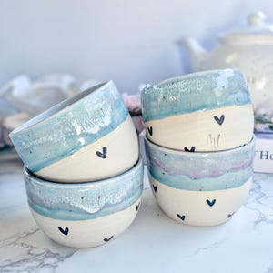 Könnte beinhalten: Drei Keramikschalen mit cremefarbener Basis und einem gesprenkelten, blau-violett glasierten Rand. Jede Schale ist mit kleinen, handgemalten schwarzen Herzmotiven verziert, die über die Außenseite verteilt sind. Die Schalen sind gestapelt.