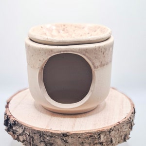 Könnte beinhalten: Ein weißer Keramik-Ölbrenner mit gesprenkelter Glasur steht auf einem Holzständer mit drei Beinen. Der Brenner hat eine runde Öffnung in der Mitte und eine kleinere Schale oben.
