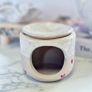 May include: A ceramic wax warmer featuring a two-tone design with a speckled white upper section and a solid beige lower section. The base has a large circular opening for a tea light and is decorated with small, hand-painted red heart motifs.