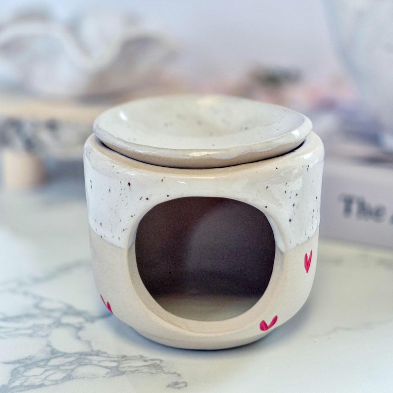 May include: A ceramic wax warmer featuring a two-tone design with a speckled white upper section and a solid beige lower section. The base has a large circular opening for a tea light and is decorated with small, hand-painted red heart motifs.