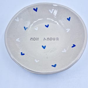 May include: A small, round, off-white ceramic bowl with a slightly uneven rim. The bowl is decorated with blue and white heart shapes around the edge and the words "MON AMOUR" in the center.