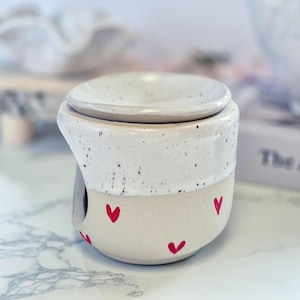 May include: A ceramic wax warmer featuring a two-tone design with a speckled white upper section and a smooth cream-colored base. The lower portion is decorated with small, hand-painted red hearts. It includes a removable dish on top.