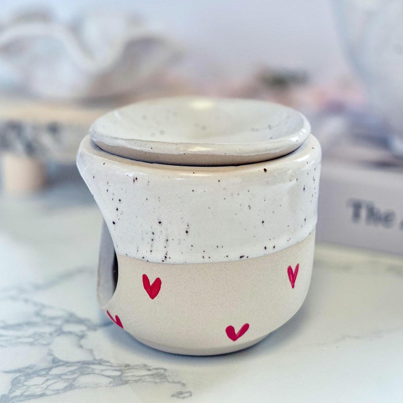 May include: A ceramic wax warmer featuring a two-tone design with a speckled white upper section and a smooth cream-colored base. The lower portion is decorated with small, hand-painted red hearts. It includes a removable dish on top.