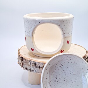 May include: A ceramic wax warmer with a speckled white upper section and a cream-colored base featuring small red heart motifs. The piece has a large circular opening on the front and is accompanied by a matching speckled ceramic dish for wax melts.