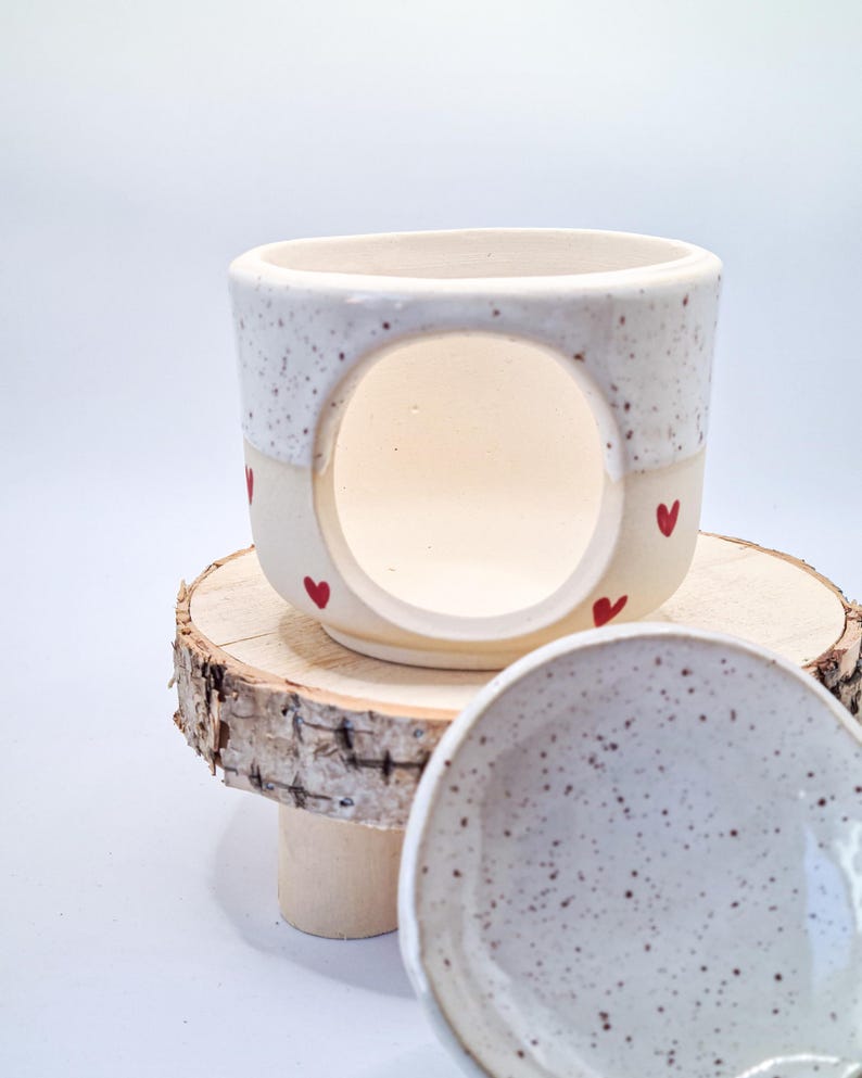 May include: A ceramic wax warmer with a speckled white upper section and a cream-colored base featuring small red heart motifs. The piece has a large circular opening on the front and is accompanied by a matching speckled ceramic dish for wax melts.