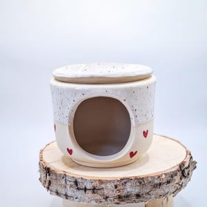 May include: A ceramic wax warmer featuring a speckled white upper section and a smooth cream-colored base. The cylindrical body has a large circular opening in the front and is decorated with small, hand-painted red hearts around the exterior.