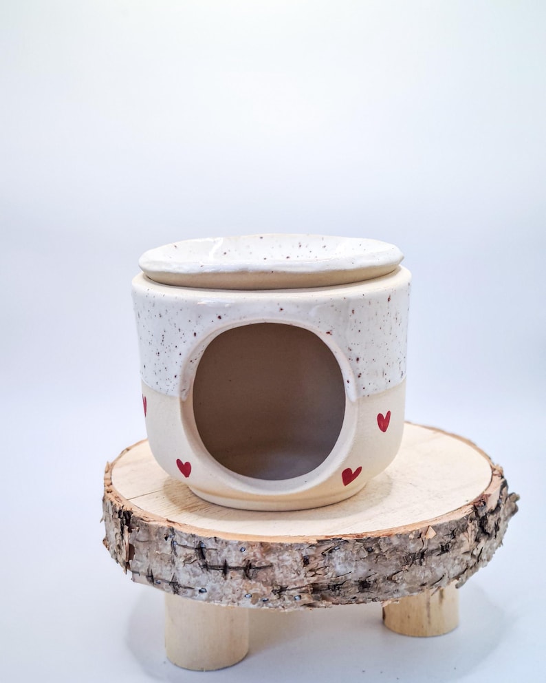 May include: A ceramic wax warmer featuring a speckled white upper section and a smooth cream-colored base. The cylindrical body has a large circular opening in the front and is decorated with small, hand-painted red hearts around the exterior.