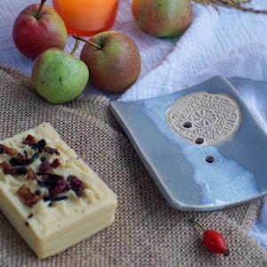 Könnte beinhalten: Eine hellblaue Keramik-Seifenschale mit einem dekorativen Design und einem Abflussloch. Die Schale liegt auf einem weißen Tuch mit einer Seife und Äpfeln im Hintergrund.