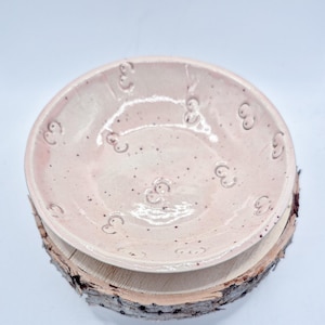 May include: A handmade, light pink ceramic bowl with a speckled finish and embossed decorative elements. The bowl is placed on a natural wood slice, creating a rustic and artistic presentation. The bowl's design is simple yet elegant.