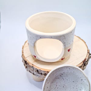 May include: A ceramic wax warmer with a speckled white upper section and a smooth cream-colored base featuring small red heart accents. The cylindrical body has a large circular opening on the front, and a separate speckled white ceramic dish is included.