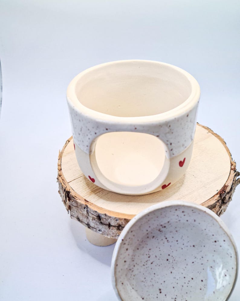 May include: A ceramic wax warmer with a speckled white upper section and a smooth cream-colored base featuring small red heart accents. The cylindrical body has a large circular opening on the front, and a separate speckled white ceramic dish is included.