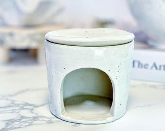 May include: A cylindrical ceramic oil burner with a speckled off-white glaze. The piece features a wide arched opening at the base for a tea light and a removable circular dish on top for wax melts or essential oils, showcasing a smooth finish.