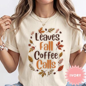May include: An ivory t-shirt featuring the text "Leaves Fall Coffee Calls" in brown and orange lettering. The design is surrounded by a circular arrangement of colorful autumn leaves and small coffee beans, creating a seasonal graphic print.
