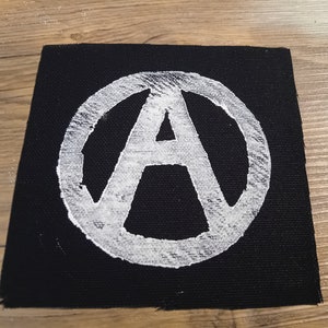 May include: A white anarchy symbol, an A inside a circle, is ironed onto a black fabric patch.