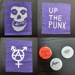 May include: Four purple fabric patches with white designs. The patches feature a skull, the words "UP THE PUNX", a transgender symbol with an anarchy symbol, and a Green Day logo. There are also three round buttons with designs. One button is red with the words "down 2 clown", one is white with the words "Xray Spex", and one is white with a Green Day logo.