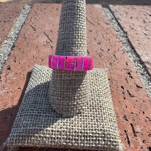 May include: A vibrant pink ring with a rectangular design, set on a silver band. The ring is displayed on a burlap ring holder, set against a brick background. The ring's bright color makes it a standout accessory.