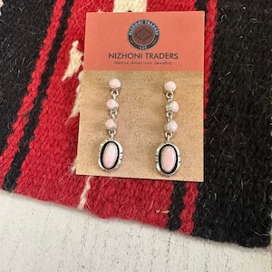 May include: A pair of pink oval stone dangle earrings with silver accents. The earrings are displayed on a card that reads "Nizhoni Traders, Native American Jewelry".