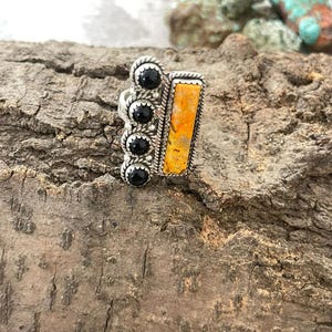 Op de afbeelding: Een zilveren ring met een rechthoekige, oranje-gele steen en drie zwarte stenen. De ring heeft een sierlijk ontwerp met een getextureerde uitstraling. De ring wordt gepresenteerd op een stuk hout.
