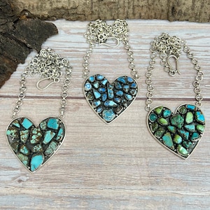 May include: Three heart-shaped necklaces with silver chains. Each pendant is filled with turquoise-colored stones. The necklaces are displayed on a light wooden surface with a piece of bark in the background.