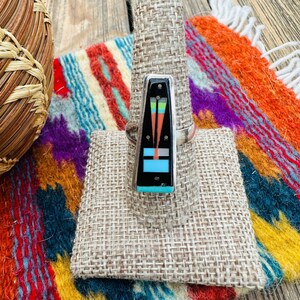 May include: A silver ring with a rectangular black inlay featuring a geometric design in turquoise, orange, and green. The ring is displayed on a burlap-covered stand, set against a backdrop of a colorful woven textile.