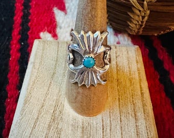 May include: A silver ring with a turquoise stone center, displayed on a wooden ring holder. The ring has a detailed, ornate design with a floral or sunburst pattern. The background includes a red, black, and white patterned textile and a woven basket.