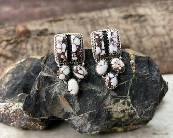 May include: A pair of silver-toned earrings with a rectangular top and three oval-shaped stones. The earrings feature white and brown speckled stones with a black stripe. The earrings are displayed on a dark gray rock.
