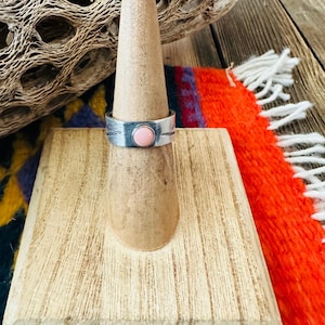 May include: A silver ring with a pink stone set in the center. The ring is on a wooden ring display stand.