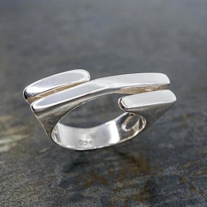 May include: A sterling silver ring with a modern, abstract design. The ring features a smooth, polished finish and a unique shape with three raised, rectangular elements. The ring is set against a dark gray background.
