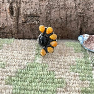 Op de afbeelding: Een zilveren ring met een centrale zwarte steen, omringd door een cluster van oranje-gele stenen. De ring wordt gepresenteerd tegen een getextureerde achtergrond, die het ontwerp en het kleurcontrast van het sieraad benadrukt.
