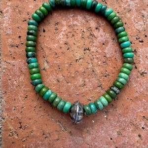May include: A beaded bracelet featuring a mix of green hues. The bracelet is made of small, round beads and includes a silver-colored, spherical accent bead. The bracelet is displayed on a textured, reddish-brown surface.