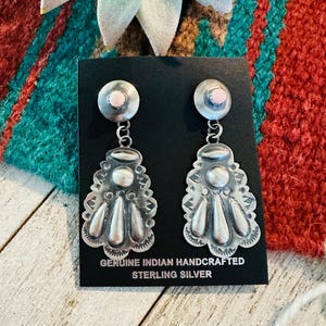 May include: A pair of sterling silver earrings with a detailed, handcrafted design. Each earring features a round top with a pink stone, connected to a decorative pendant with a scalloped edge and teardrop shapes. The text "GENUINE INDIAN HANDCRAFTED STERLING SILVER" is visible.