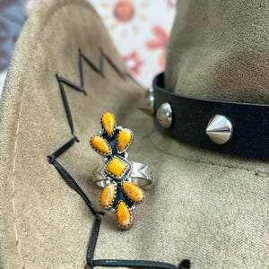 Op de afbeelding: Een zilveren ring met een cluster van oranje edelstenen. De ring ligt op een bruin cowboyhoed met een zwarte leren band en zilveren studs. Het ontwerp van de ring toont een bloemvormige opstelling van de oranje stenen.