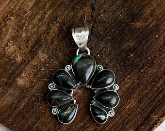May include: A silver pendant with a horseshoe-shaped design. The pendant features a large, teardrop-shaped black stone in the center, surrounded by smaller black stones. The stones have a marbled appearance. The pendant has a turquoise stone at the top.