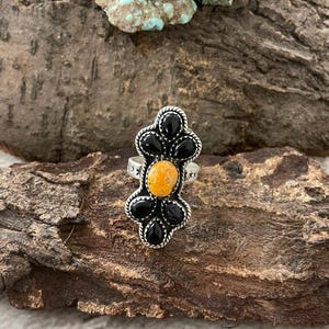 Op de afbeelding: Een zilveren ring met een centrale oranje steen, omringd door zwarte, druppelvormige stenen. De ring heeft een bloemmotief en ligt op een getextureerd houten oppervlak. De ringband heeft stervormige details.