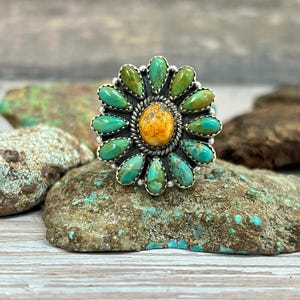 Op de afbeelding: Een zilveren ring met een bloemmotief. Het heeft een centrale oranje steen, omringd door turquoise en groene stenen. De ring wordt gepresenteerd op een getextureerd stenen oppervlak, dat de details benadrukt.