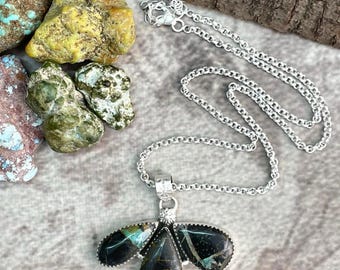 May include: A silver necklace with a pendant featuring a dark, teardrop-shaped stone with green and blue accents. The pendant has two wing-shaped stones on either side. The necklace is displayed on a textured surface.