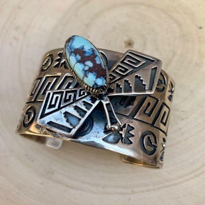 Może przedstawiać: Srebrna bransoleta z turkusem i geometrycznymi wzorami. Owalny turkus ma brązowe wzory macierzy. Bransoleta ma wytrawione wzory i prostokątny element akcentujący. Mankiety są prezentowane na drewnianej powierzchni.