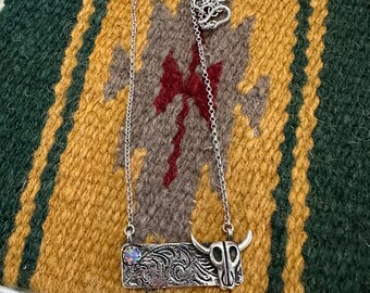 May include: A silver necklace with a rectangular pendant featuring an ornate design and a small opal stone. The pendant is attached to a silver bull skull charm.
