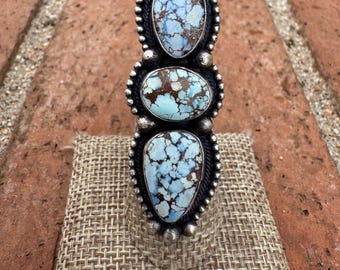 May include: A silver ring featuring three oval-shaped, light blue turquoise stones with brown matrix patterns. The stones are set in a silver setting with a beaded border. The ring is displayed on a textured, tan surface.