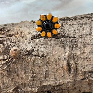 May include: A silver-toned ring with a flower design. The ring features a black center stone surrounded by orange-yellow stones. The ring is set on a piece of wood.