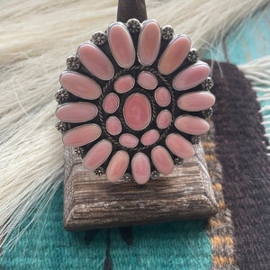 May include: A silver ring with a pink abalone shell inlay. The shell is cut into a flower shape with a central oval surrounded by smaller ovals.