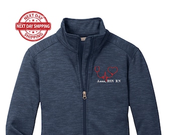May include: A navy blue fleece jacket with a zipper closure and two pockets. The jacket has a red embroidered stethoscope and heart with the text "Anna, BSN RN".