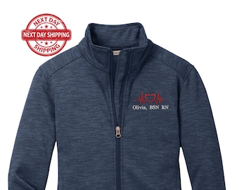 May include: A navy blue women's zip-up jacket with a red heart and EKG line embroidered on the left chest. The text "Olivia, BSN RN" is embroidered below the heart.