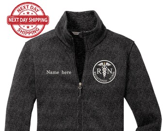 May include: A dark gray fleece zip-up jacket with a white embroidered logo that says "Registered Nurse" and a caduceus symbol. The jacket has a zipper closure and two pockets.